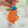 Hand holding a small orange crocheted character keychain with a blue bow against a blurred background.